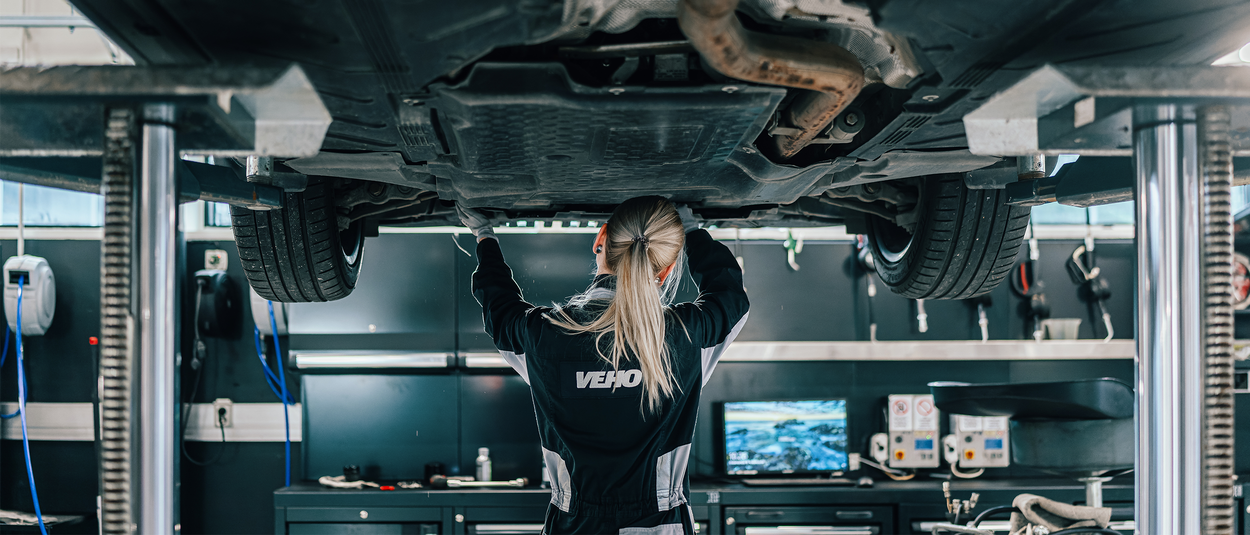 Vehon Mercedes-mekaanikko selkä kameraan auton alla kiinnittämässä moottorin pohjamuovia.