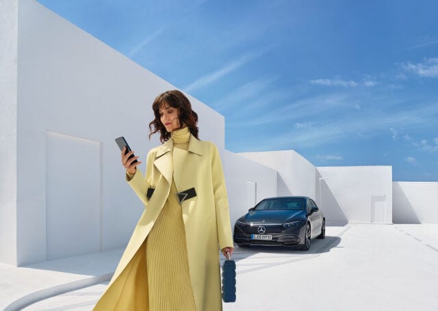 Nuori tyylikkäästi pukeutunut nainen katsoo puhelinta, taustalla tumma Marcedes benz auto